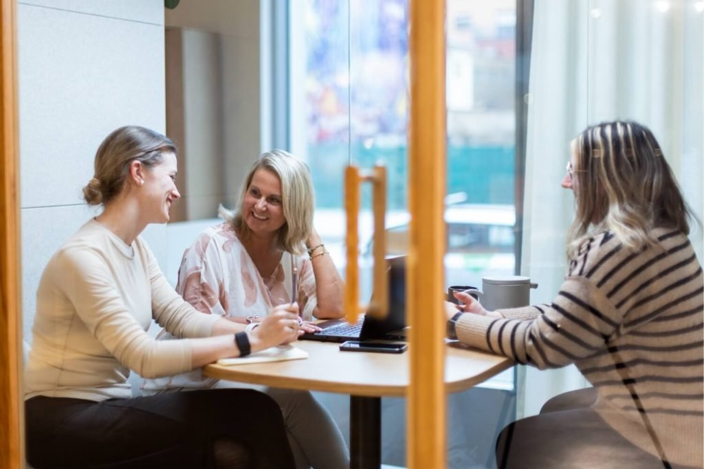 Tres mujeres sonríen y conversan durante una reunión alrededor de una mesa con un portátil y tazas. Están en el interior de una cabina insonorizada Chat4 GEN4.