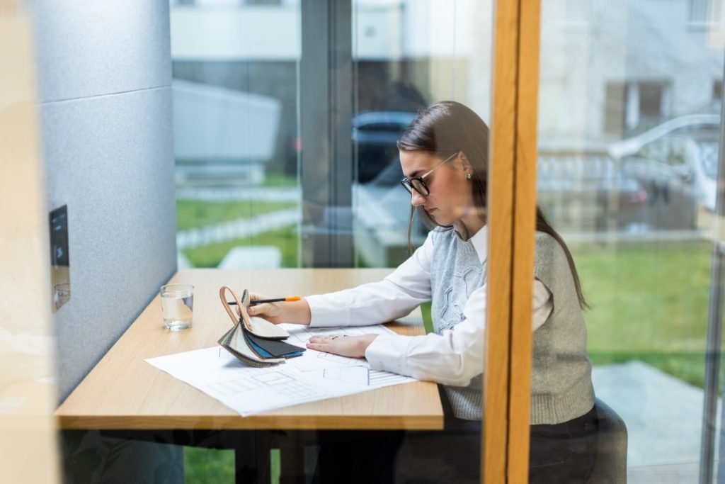 Mujer con gafas dibujando sobre planos arquitectónicos junto a muestras de materiales dentro de una cabina acústica Focus GEN4