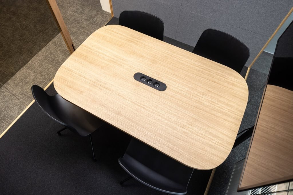 Mesa de reuniones de madera con cuatro sillas negras y tomas de corriente integradas en el centro. Detalles del interior de una cabina acústica Summit4 GEN4