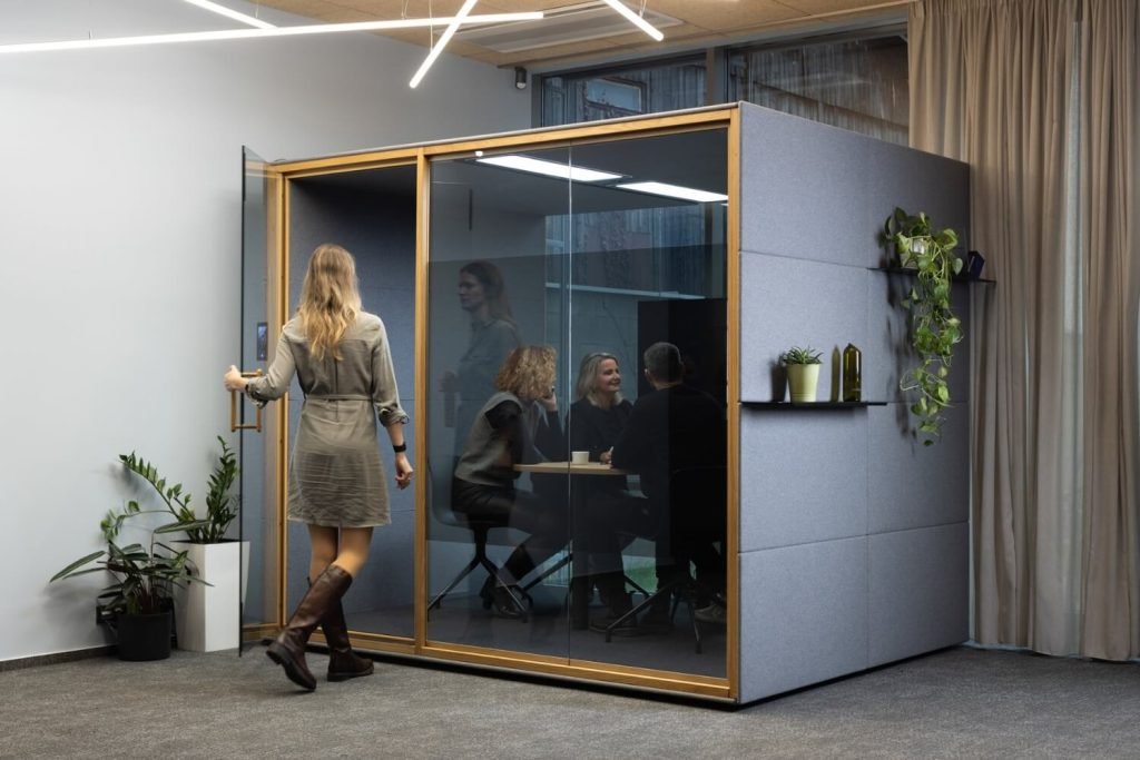Una mujer entra en una cabina acústica Summit4 GEN4 de reuniones acristalada donde tres personas conversan alrededor de una mesa