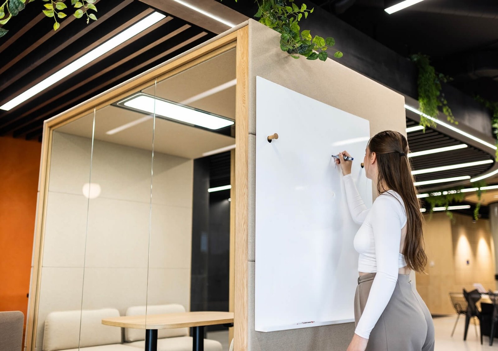 Una mujer escribe en una pizarra blanca de una Cabina Acústica Chat2 dentro de una oficina moderna. Workplace Trends