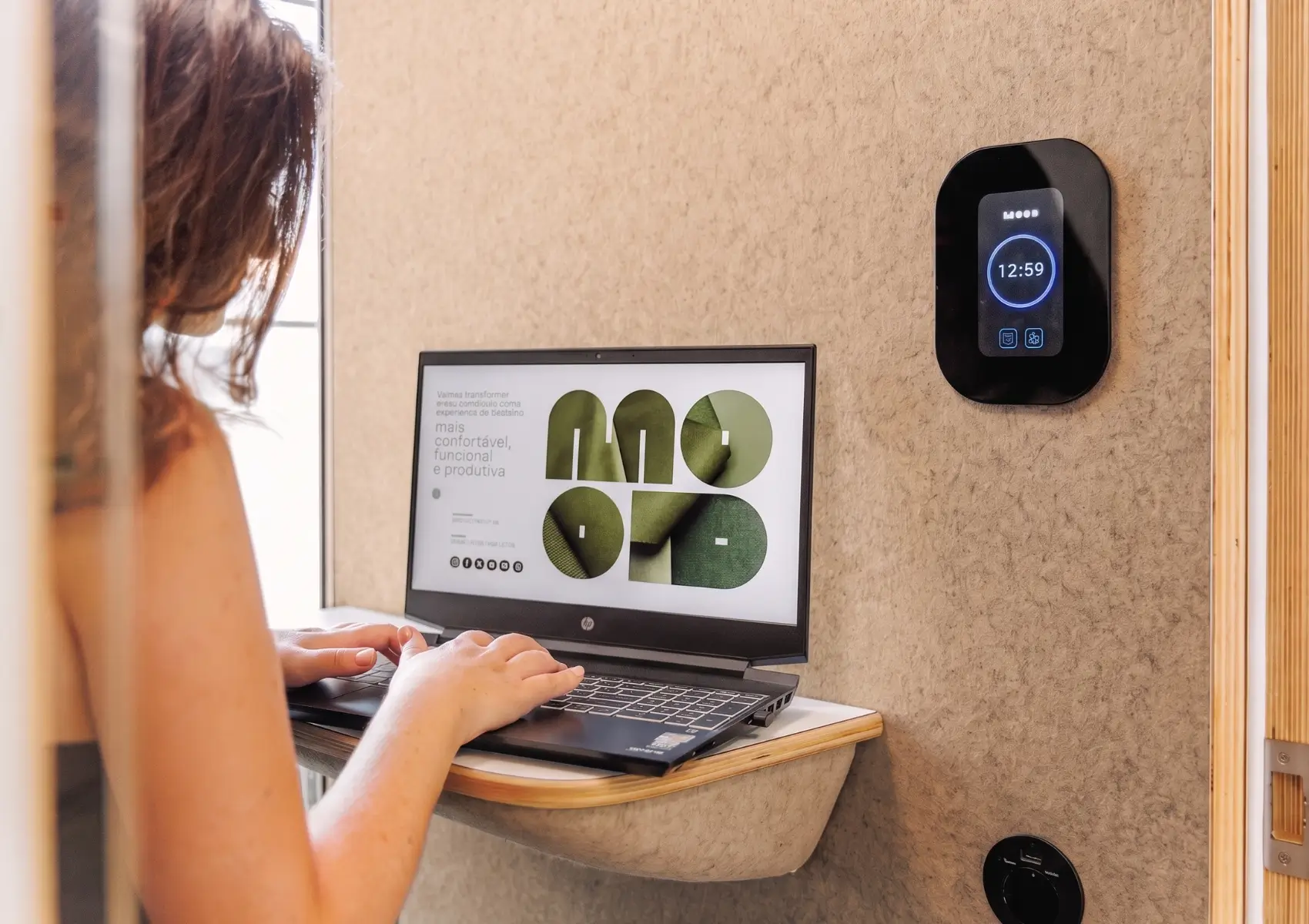 Persona trabajando con un portátil en un escritorio plegable, junto a controles inteligentes en la pared de una cabina acústica MOOD Phone. Pude consultar nuestra página de Preguntas Frecuentes para más dudas.